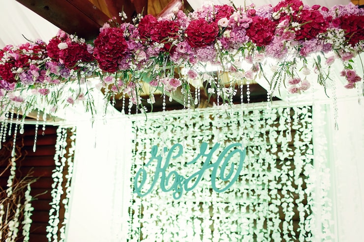 A lush, elegant wedding or special event backdrop featuring an overhead floral arrangement of crimson and pink hydrangeas, dangling crystal garlands, and a personalized wooden initial monogram (H&H) set against a delicate, illuminated hanging garland curtain.