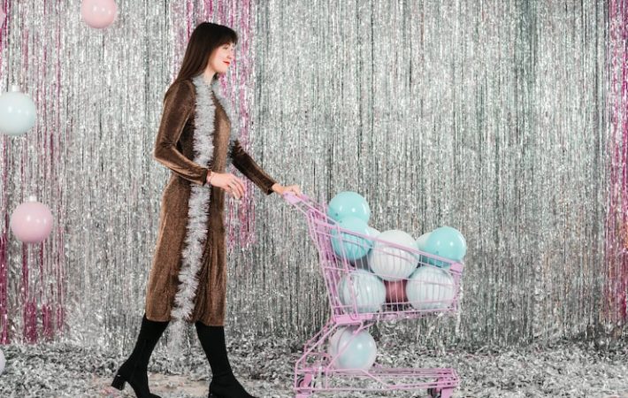 A woman posing in front of a floor-to-ceiling silver foil fringe backdrop while pushing a miniature shopping cart filled with pastel balloons, illustrating a sparkly, vibrant party theme ideal for a milestone birthday or corporate event.
