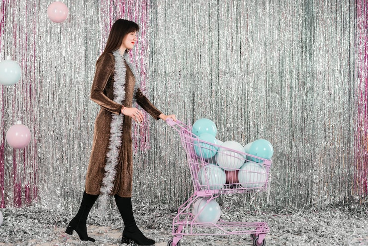 A woman posing in front of a floor-to-ceiling silver foil fringe backdrop while pushing a miniature shopping cart filled with pastel balloons, illustrating a sparkly, vibrant party theme ideal for a milestone birthday or corporate event.