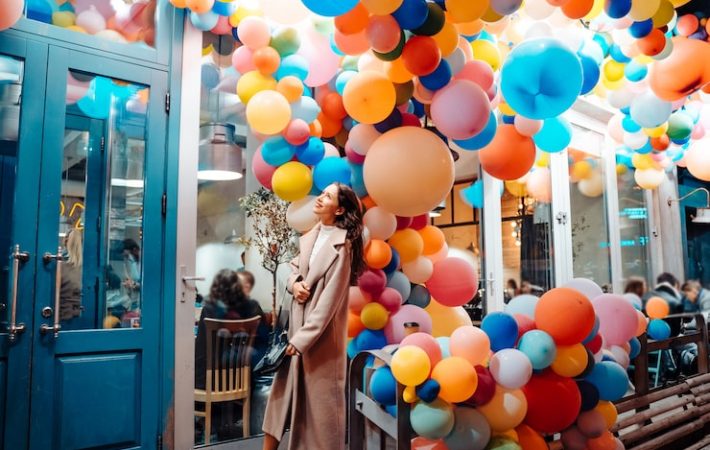 A woman in a long beige coat stands admiring a dense, colorful overhead installation of brightly colored balloons in shades of orange, yellow, pink, and blue, spilling out from the entrance of a building with a bright blue door frame.