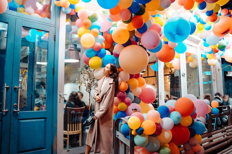 A woman in a long beige coat stands admiring a dense, colorful overhead installation of brightly colored balloons in shades of orange, yellow, pink, and blue, spilling out from the entrance of a building with a bright blue door frame.