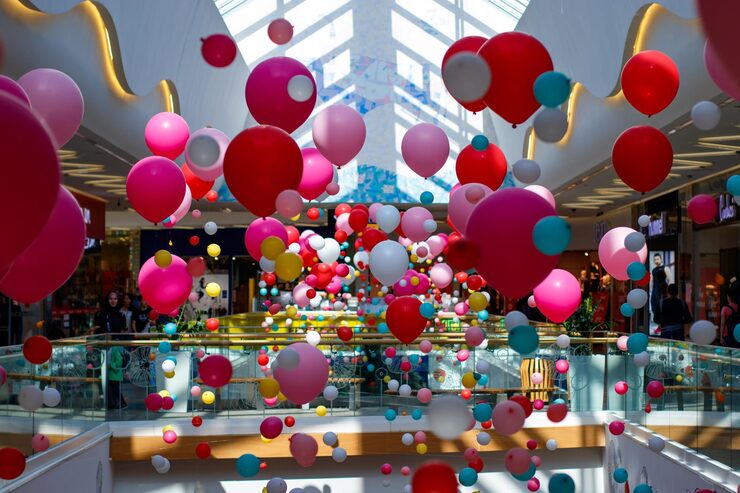 A large, open, multi-story indoor venue, possibly a shopping mall, is decorated with hundreds of loose balloons in various shades of pink, white, red, and blue, floating and hanging at different heights above the railings of the upper levels.