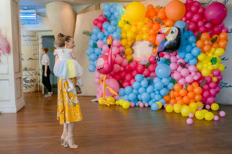 A bright and vibrant balloon arch or wall, featuring an array of blue, pink, yellow, and orange balloons, including novelty balloons shaped like a flamingo and a toucan, serves as a backdrop for a woman in a yellow floral skirt holding a small child in a white and yellow dress. How to Choose Balloon Arches That Suit Your Toronto Venue