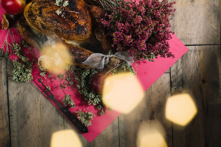 A warm, top-down view of a roasted whole chicken or turkey resting on a vibrant magenta cloth over a rustic wooden table. It is garnished with fresh herbs and a cluster of deep pink/burgundy heather flowers. Apples and a fig are visible nearby, and the scene is accented by large, soft hexagon-shaped light bokeh in the foreground.