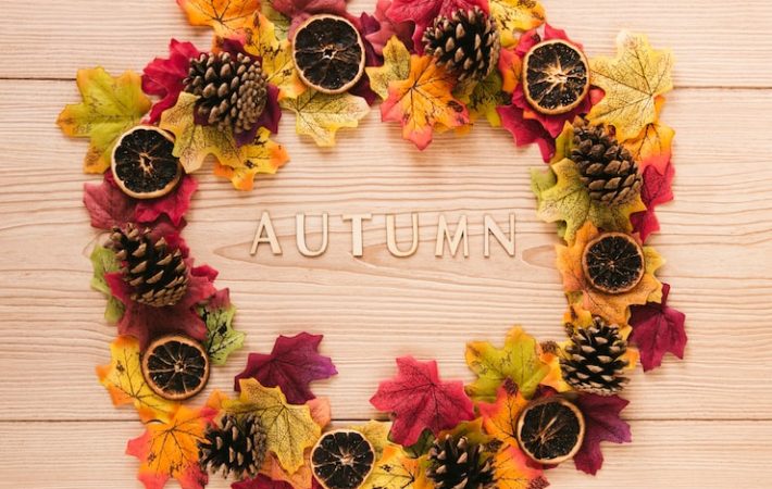 A flat lay of a circular autumn wreath resting on a light wood plank background. The wreath is composed of vibrant red, orange, and yellow faux maple leaves, interspersed with pinecones and several slices of dried citrus. The wooden letters spelling "AUTUMN" are positioned horizontally in the center of the wreath. Fall seasonal flower theme ideas for toronto parties