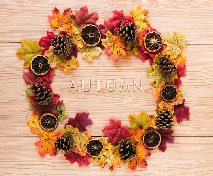 A flat lay of a circular autumn wreath resting on a light wood plank background. The wreath is composed of vibrant red, orange, and yellow faux maple leaves, interspersed with pinecones and several slices of dried citrus. The wooden letters spelling "AUTUMN" are positioned horizontally in the center of the wreath. Fall seasonal flower theme ideas for toronto parties