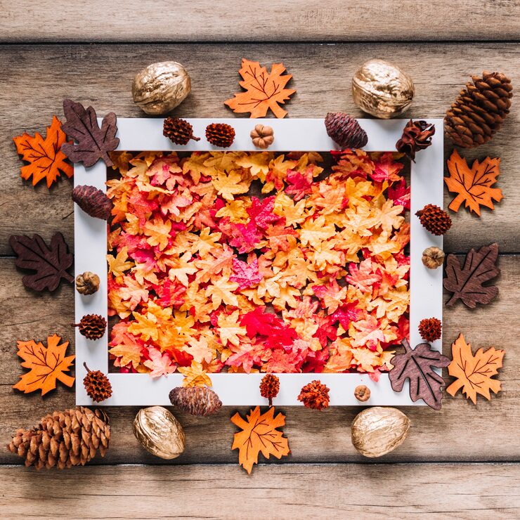 A flat lay of a square white picture frame filled with a dense, colorful mix of faux maple leaves in hues of red, orange, and yellow. The frame is centered on a rustic dark wood surface and surrounded by assorted fall decor, including scattered pinecones, small decorative gourds, dark brown wooden leaf cutouts, and several shiny gold-painted walnuts.