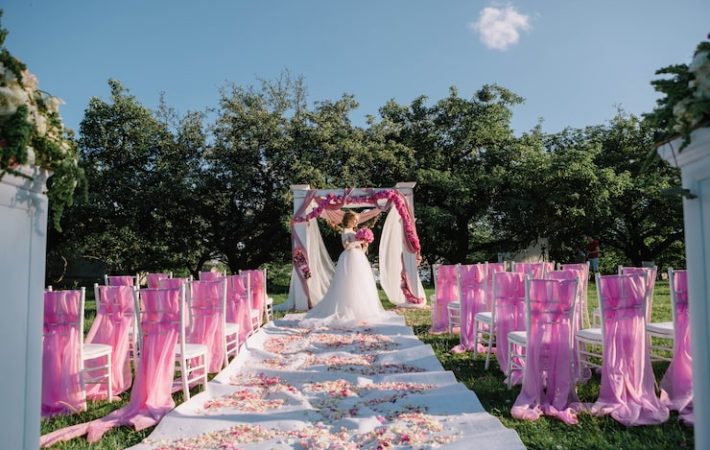 A bride stands under a wedding arch rental covered in pink and white fabric and flowers, marking the ceremony space outdoors. The aisle is lined with white chairs draped in bright pink chiffon and scattered rose petals, illustrating a vibrant floral installation for a wedding in Toronto. Floral Installations for Weddings in Toronto