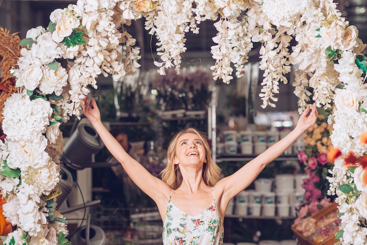 A smiling woman with her arms raised stands under a large, lush white and cream floral wedding arch. The abundance of white roses and hanging wisteria-like flowers highlights a dramatic, romantic Toronto wedding flower package or installation.