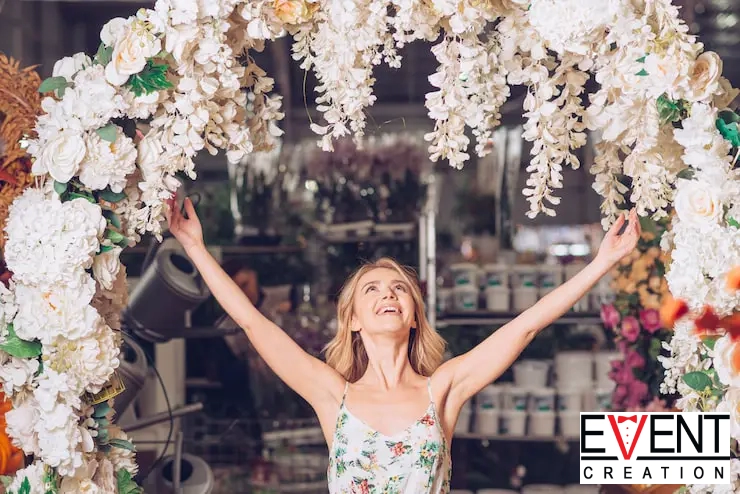A smiling woman with her arms raised stands under a large floral wedding arch.