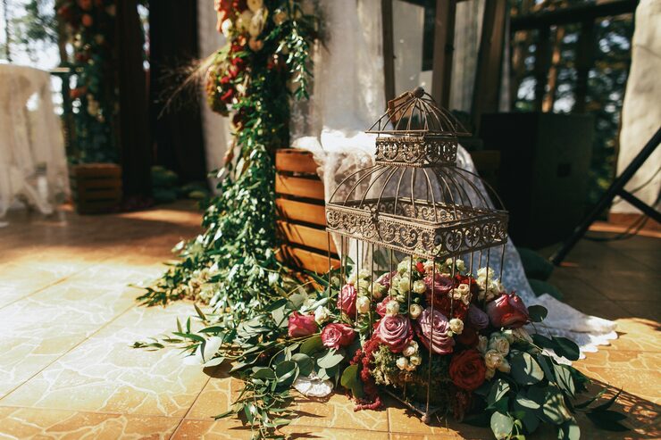 A close-up of vintage-style wedding decor rentals Toronto featuring an antique bronze birdcage overflowing with deep burgundy and blush roses, surrounded by vibrant green ivy and eucalyptus, set on a tiled floor for an outdoor or rustic ceremony.
