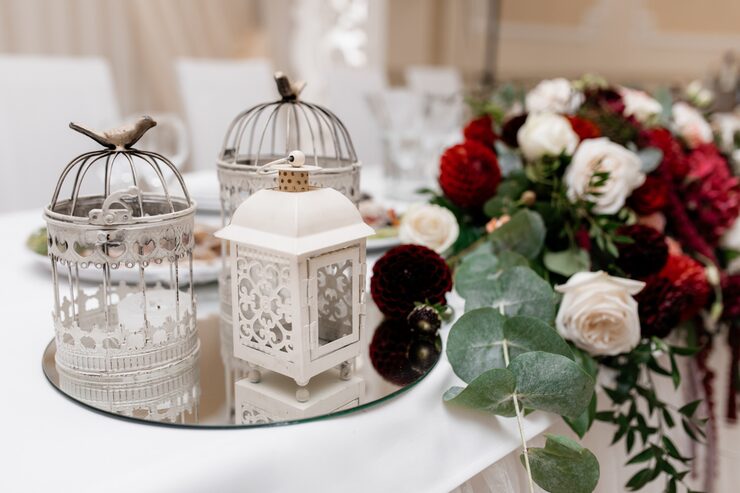 An elegant close-up of a wedding table centerpiece featuring vintage-style white metal birdcages and a small lantern resting on a circular mirrored tray. To the right is a vibrant floral arrangement with deep red dahlias, white roses, and trailing eucalyptus and green foliage.