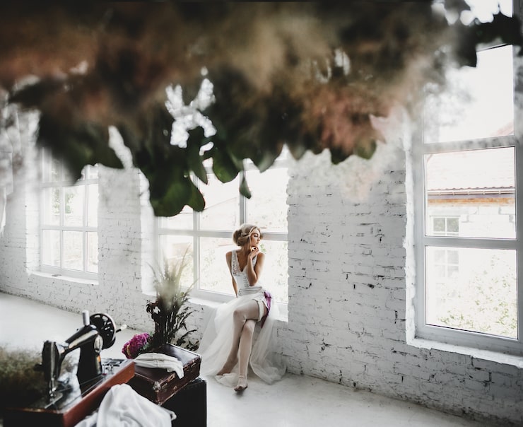 A bridal portrait taken in a bright, industrial-style room with white brick walls and large windows. The bride is seated gracefully on a wide windowsill, wearing a delicate white gown and heels. The foreground includes blurred, hanging foliage and an antique sewing machine on a wooden trunk, suggesting a romantic, vintage aesthetic.