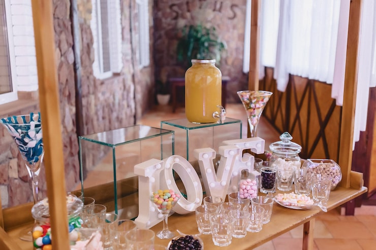 A colorful candy and dessert bar setup on a rustic wooden cart at a wedding reception. The display features a large dispenser of yellow beverage, various glass jars filled with colorful candies and sweets, clear drinking glasses, and large light-up marquee letters spelling "LOVE" as a centerpiece.