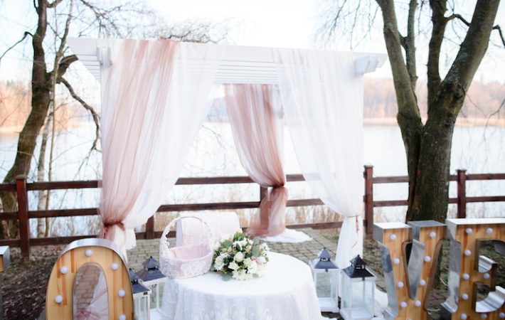 An outdoor wedding ceremony arch or pergola set up near a body of water, possibly for an engagement shoot or small reception. The white wooden structure is draped with flowing white and sheer, dusty-rose colored fabric. In the center, a small round table with a lace cloth holds a bouquet of white and green flowers and a decorative woven basket. Large light-up marquee letters spelling "LOVE" are visible on the ground to the right.