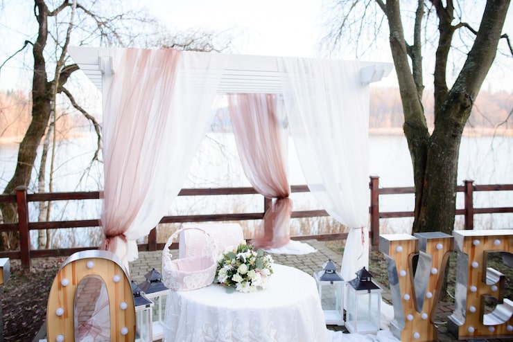 An outdoor wedding ceremony arch or pergola set up near a body of water, possibly for an engagement shoot or small reception. The white wooden structure is draped with flowing white and sheer, dusty-rose colored fabric. In the center, a small round table with a lace cloth holds a bouquet of white and green flowers and a decorative woven basket. Large light-up marquee letters spelling "LOVE" are visible on the ground to the right.