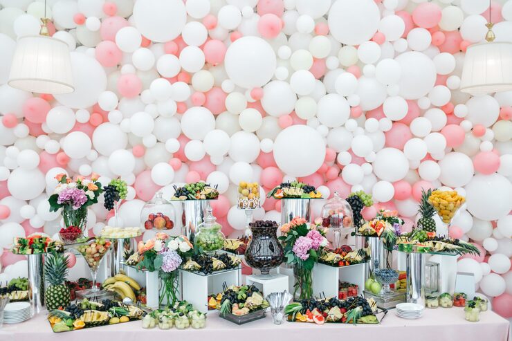 A full wall backdrop featuring an dense pattern of white and pale pink balloons of various sizes, with an elaborate table of fresh fruit and floral centerpieces in front.
